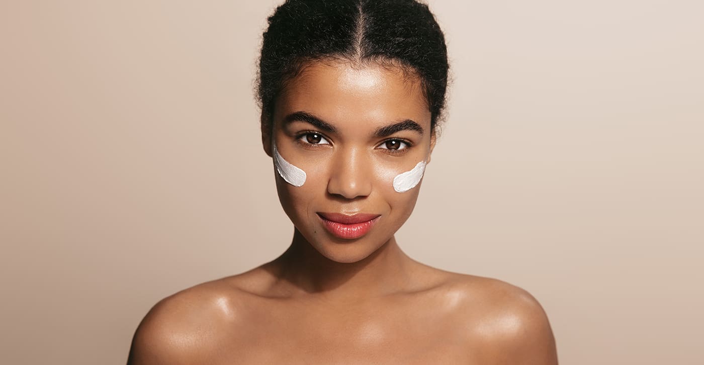 Vertical portrait of woman with healthy face using facial cream, moisturizer, skincare product on cheek, using lotion or daily care for better skin tone, brown background